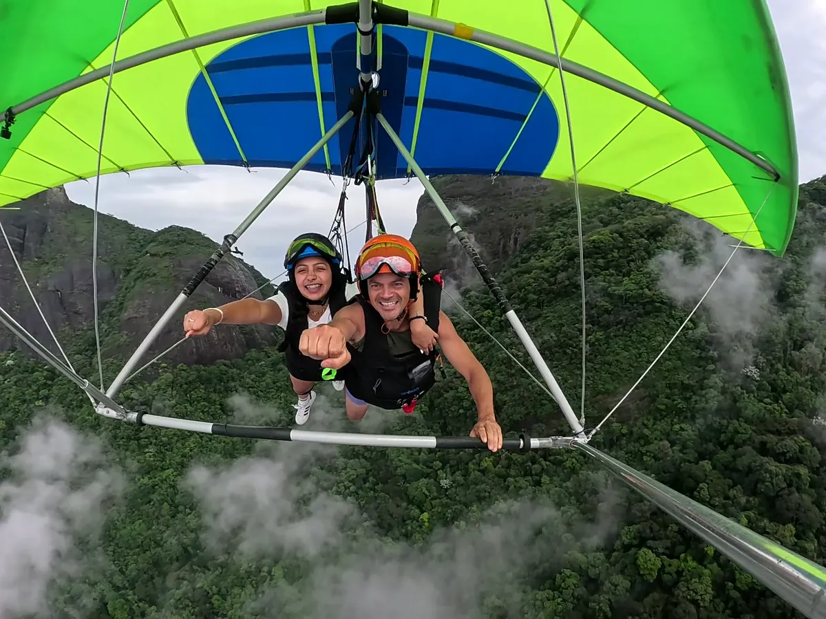 Passageira e piloto sorrindo em selfie GoPro durante voo de asa-delta sobre a floresta e as nuvens do Rio de Janeiro